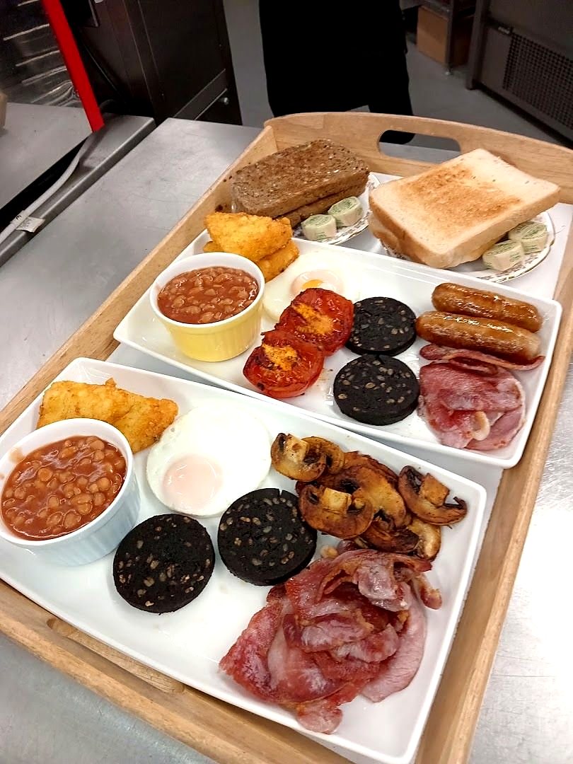 Two full breakfasts on a wooden tray: bacon, sausages, black pudding, beans, mushrooms, eggs, tomatoes, hash browns and toast