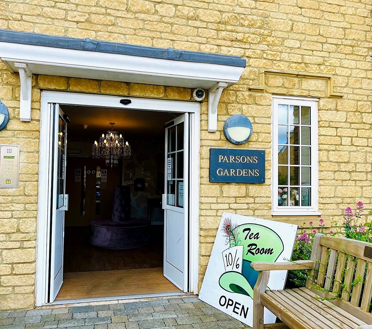 The tea room’s entrance in a honey-coloured Cotswold stone building: open white double doors, a navy Parsons Gardens plaque, a wooden bench, and a sign shaped like a green top hat with a 10/6 card and the word OPEN